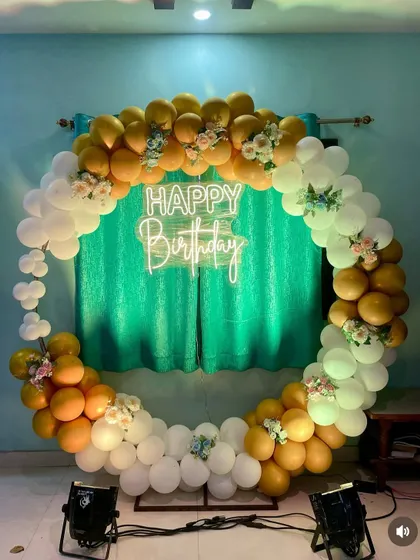 A simple yet elegant birthday decoration with a circular balloon frame. The yellow and white balloons are accented with small floral bouquets and a neon sign.