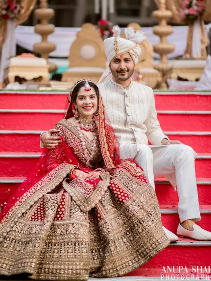A classic wedding portrait of the bride and groom, seated on the steps of the mandap. They look regal and happy in their traditional red and white wedding attire.