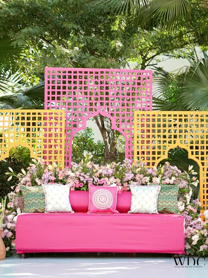 A beautiful seating area from our 'Serene Sage' Mehendi. The vibrant pink sofa is set against a backdrop of yellow and pink jali panels, surrounded by lush florals, creating a perfect blend of color and tradition.