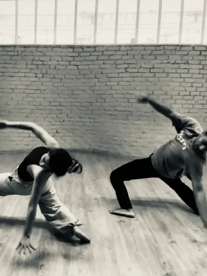 Two dancers in a powerful, grounded pose during a rehearsal for 'Dream Baby Dream'. The black and white filter highlights the raw physicality of the performance.