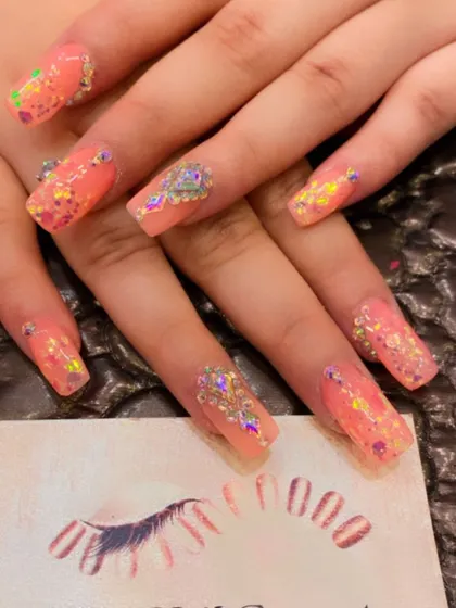 A bright and sparkling set of peach-colored square nails. The design is heavily embellished with chunky iridescent glitter and rhinestone clusters.