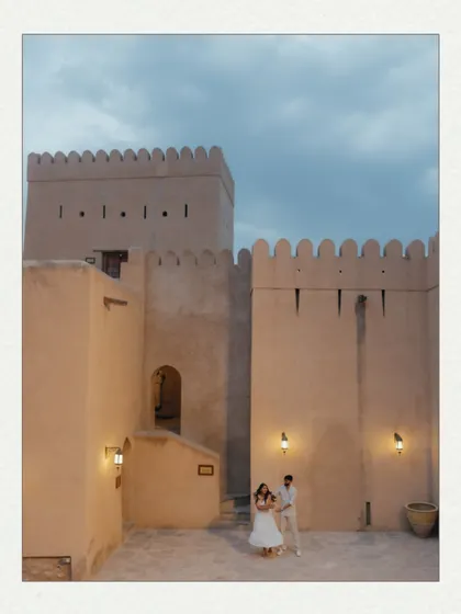 A couple dances in the courtyard of an Omani fort at dusk. The warm lights of the fort and the darkening blue sky create a magical and romantic atmosphere for this pre-wedding photo.