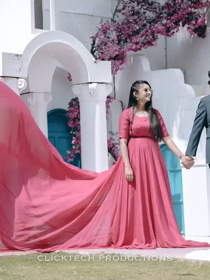 A couple poses against a Santorini-inspired white and blue backdrop, her long pink trail gown adding a beautiful flow and a pop of color to the scene.