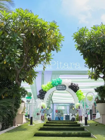 A wide view of the lush green entrance to 'Reyansh's Club', setting a sporty and exclusive tone for the tennis party.
