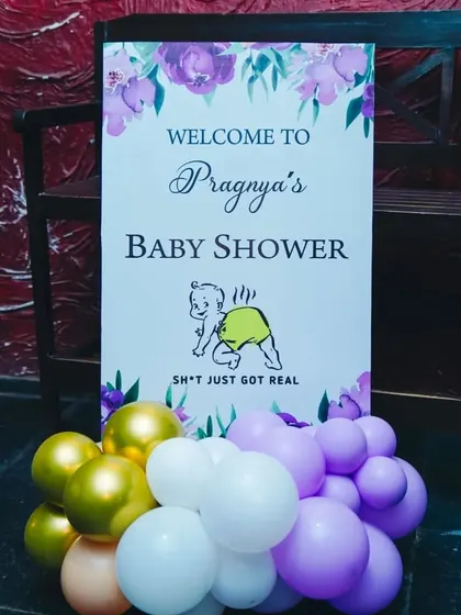 A fun and cheeky welcome sign for Pragnya's baby shower. The sign, which reads "Sh*t just got real," adds a touch of humor to the celebration, complemented by a purple and gold balloon arrangement.