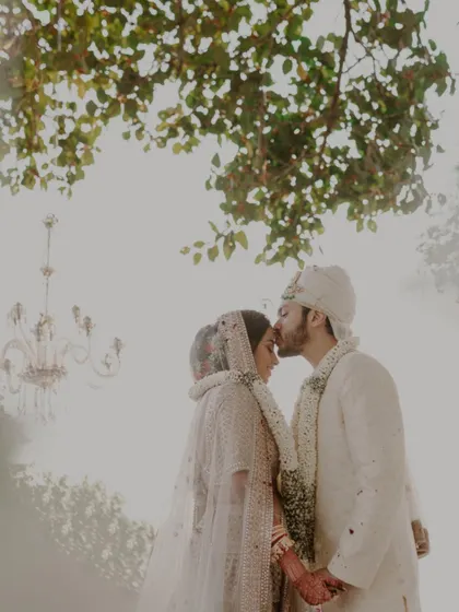 A dreamy, ethereal shot of a groom kissing his bride's forehead under a tree. The soft, hazy light and an out-of-focus chandelier create a magical and romantic wedding portrait.