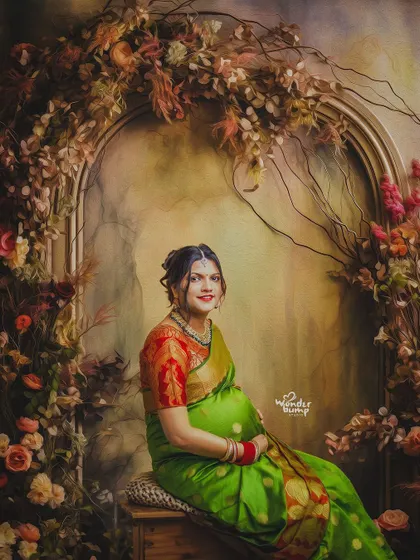 A regal portrait of a mom-to-be in a green and red saree, seated against a beautiful, ornate floral archway in our studio.