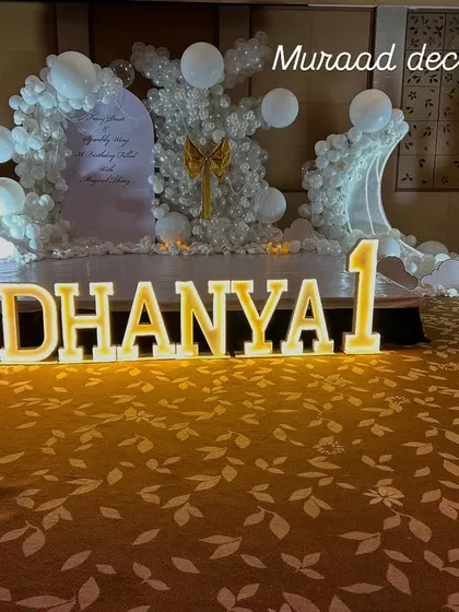 A royal first birthday for a little princess. This grand setup features large, illuminated marquee letters spelling out her name on a patterned carpet, with an all-white, castle-themed backdrop.