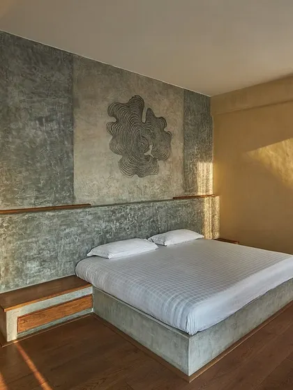 The bedroom combines raw pigmented cement walls with warm wood flooring. An abstract mural adds a touch of artistry, while the minimalist design creates a restful and serene personal space.