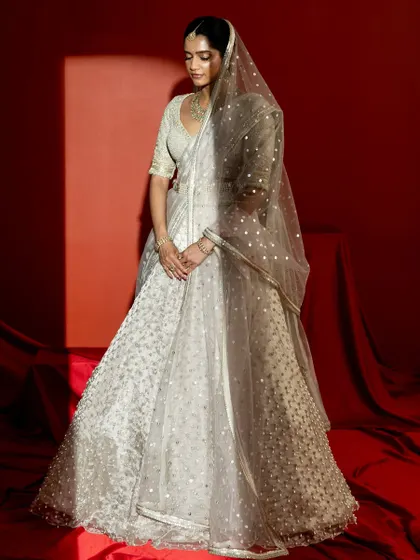 A soft, elegant pose under a spotlight, this image captures the dreamy quality of the ivory lehenga. The sheer veil adds a layer of mystique, while the intricate embroidery on the skirt and blouse subtly gleams.