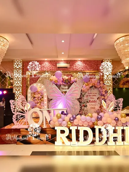 A grand butterfly-themed stage for a first birthday, featuring illuminated butterfly wings, marquee letters, and a beautiful pastel and gold balloon arrangement.