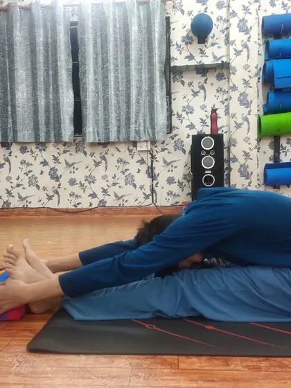 An instructor assists a student in a deep Paschimottanasana (Seated Forward Bend), using a yoga block for support. This hands-on guidance helps the student to safely explore their flexibility limits.