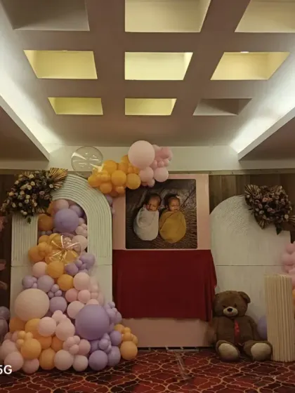 A lovely naming ceremony decoration for twins, featuring a photo of the babies at the center. The setup includes teddy bears, a picket fence, and pastel pink, purple, and orange balloons.