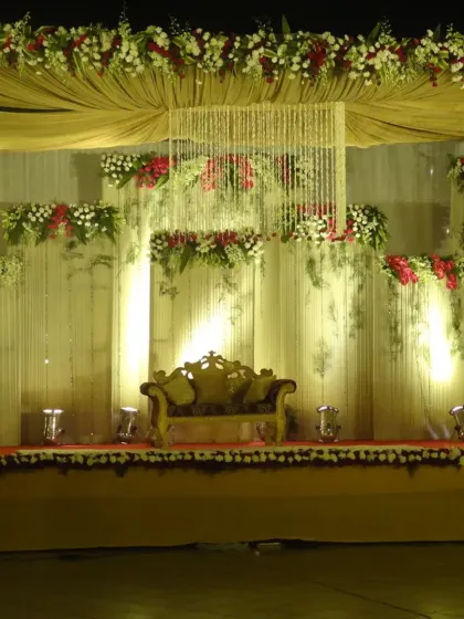 A classic stage decoration featuring a central crystal chandelier hanging from a draped ceiling. The backdrop is layered with fresh flowers in red and white, complemented by warm uplighting to create a grand and inviting focal point.
