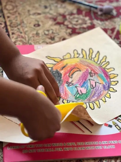 A close-up of little hands carefully cutting out a colorful sun drawing. This activity is great for practicing scissor skills.