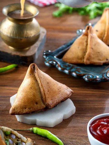A close-up shot of a crispy, golden samosa, perfectly capturing the texture of the pastry. Paired with a cup of hot chai, this image evokes the feeling of a perfect rainy day snack.