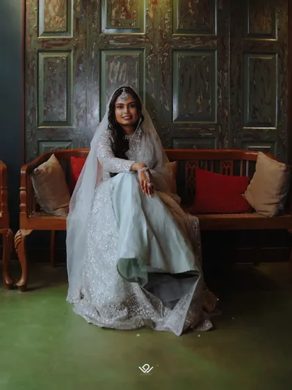 A relaxed portrait of a Muslim bride in a light blue gown, seated on a traditional wooden bench, showcasing a blend of modern style and classic settings.
