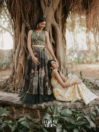 Two sisters share a laugh while sitting together under a large banyan tree. This candid portrait captures a beautiful, natural moment of sibling connection during a family celebration.