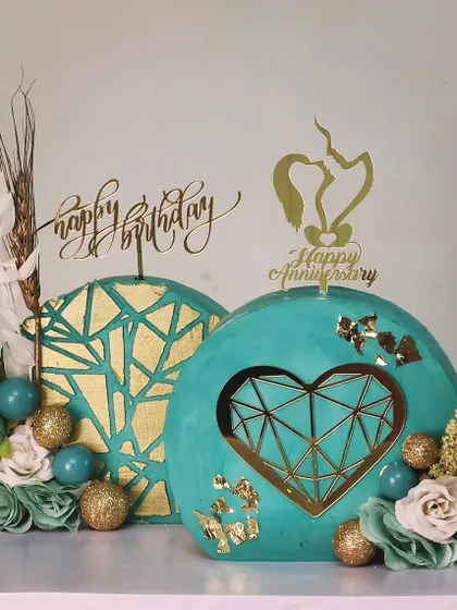 A set of two matching but distinct cakes for a joint birthday and anniversary celebration. Both feature a teal and gold color palette with modern geometric and heart-shaped designs.