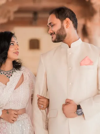 A classic walking shot through a sunlit palace corridor in Jaipur. The couple is linked arm-in-arm, sharing a look of love and admiration, dressed in elegant, complementary outfits.