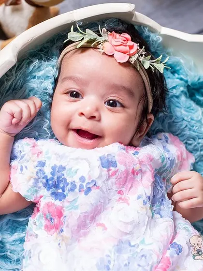 A close-up shot of a smiling baby girl in a floral dress, looking up from her cozy spot inside an eggshell prop. Her bright, happy expression is the highlight of this adorable infant picture.