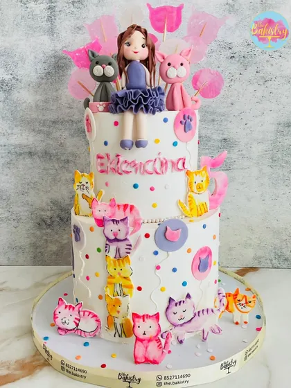 A purr-fectly adorable two-tiered cake for a cat lover. This design is covered in colorful, playful cat figures, with a sweet doll and her two favorite kitties on top.