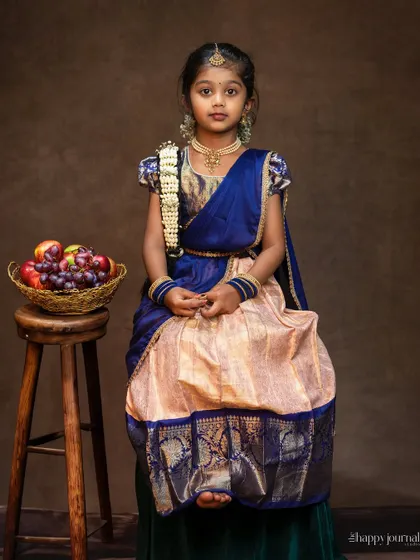A classic portrait of a girl in a traditional South Indian lehenga, sitting beside a stool with a basket of fruit. The image has a timeless, painted quality.