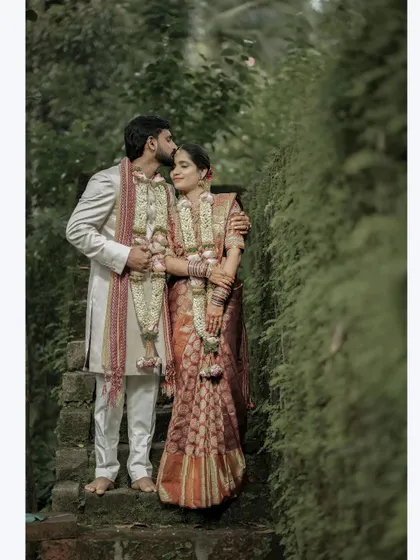 A romantic portrait of the groom kissing his bride's forehead, set against a lush green backdrop, showcasing their traditional wedding attire.