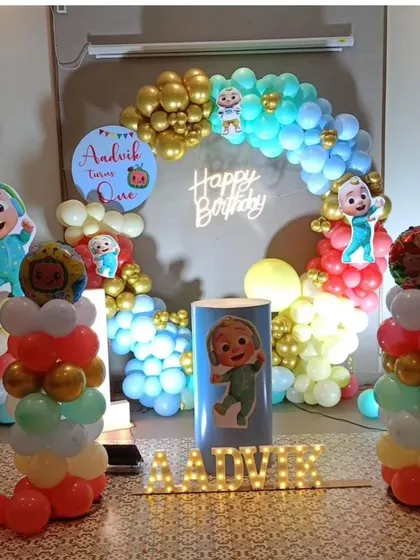 An elaborate Cocomelon first birthday setup. It includes a custom backdrop, a colorful balloon arch, multiple character cutouts, balloon pillars, and the baby's name "Aadvik" in light-up marquee letters.