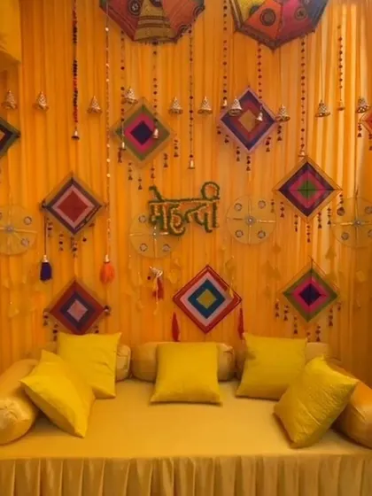A full yellow-themed Mehendi backdrop with matching seating. The decor includes hanging kites, bells, and decorative umbrellas for a cohesive and traditional look.