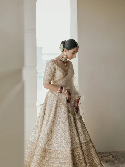 A graceful portrait of Manchal, looking down at her lehenga. The soft light from the window and her elegant posture create a serene and timeless image.