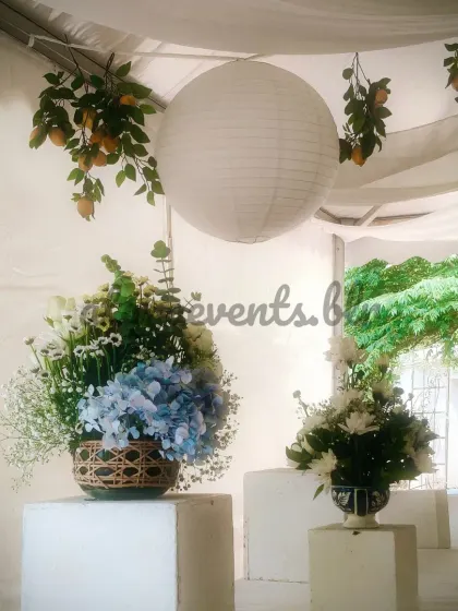 A beautiful floral centerpiece on a pedestal at a Sicilian-themed event. The arrangement of white and blue flowers is set against a backdrop of hanging lemons and white paper lanterns.