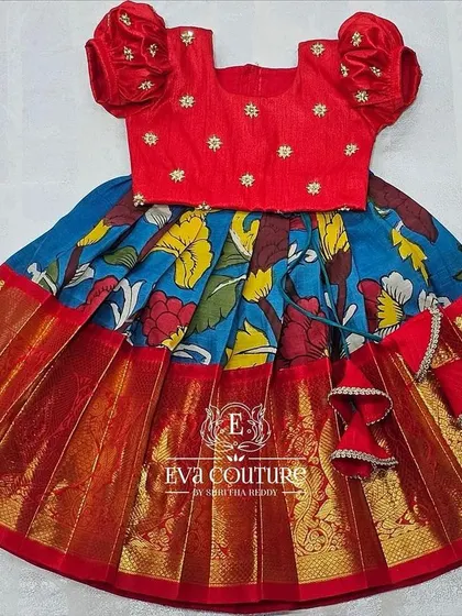 A vibrant red and blue pattu pavadai for a toddler. The blue skirt has a colorful Kalamkari-style print, paired with a simple red blouse with butti work.