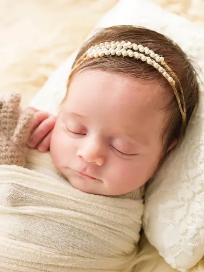 So tiny, so perfect. This close-up shows a newborn sleeping peacefully on a soft pillow, holding a tiny knitted teddy bear. Every detail is captured, from the pearl headband to her tiny fingers.