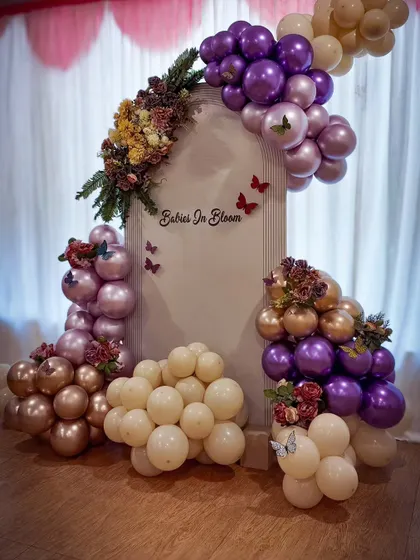 A "Babies in Bloom" setup featuring a single Chiara backdrop. The purple, gold, and cream balloons combined with floral bouquets and butterfly accents create a rich and elegant look.