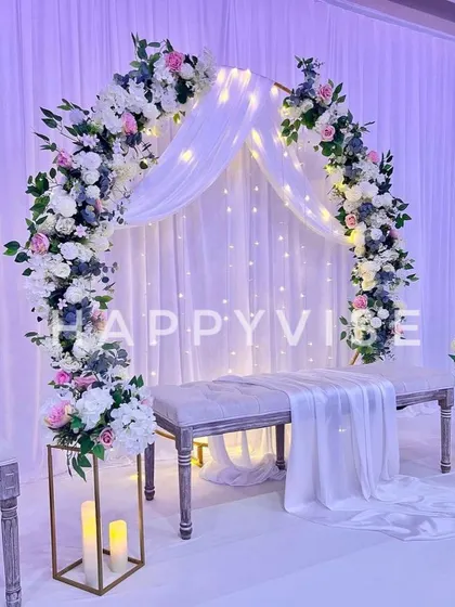 A dreamy and romantic stage with a floral arch, sheer white drapes, and twinkling fairy lights. The soft purple lighting adds a touch of magic to the scene.