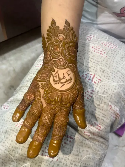 I can beautifully write names and initials in your mehendi. Here, a name in elegant Arabic script is the centerpiece of a detailed floral design on the hand.