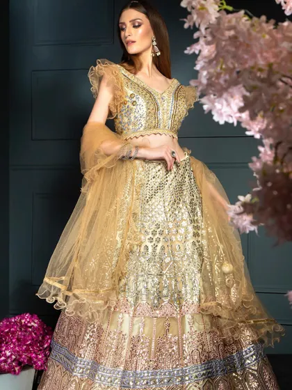 A full-length shot of a heavily embellished golden lehenga. The composition, framed by flowers, creates a soft, romantic mood perfect for bridal or festive wear collections.