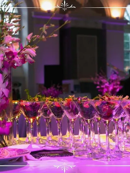 An elegant dessert and cocktail bar at a formal event. The purple ambient lighting and floral arrangements create a luxurious feel, with berry cocktails beautifully lined up for guests.