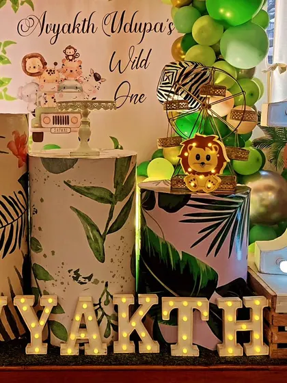A "Wild One" jungle party setup with a circular backdrop and themed plinths. The decor includes a light-up number '1' and marquee letters spelling out the birthday boy's name.