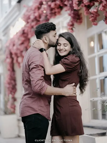 A tender moment with the groom kissing the bride's forehead, set against a backdrop of pink flowers and a cafe window.