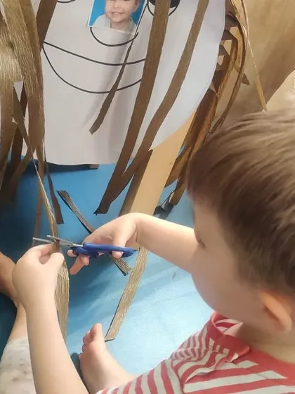 A close-up of a child carefully using child-safe scissors to give a paper face a haircut. This is a highly motivating way to practice scissor skills and hand-eye coordination.
