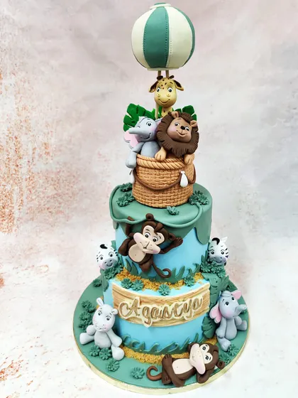 We invite you to a storybook adventure with this two-tiered animal hot air balloon cake. Meticulously crafted jungle animals, including a lion, elephant, and giraffe, are gathered in a woven basket, ready for a whimsical journey, making this a magical centerpiece for a child's birthday celebration.