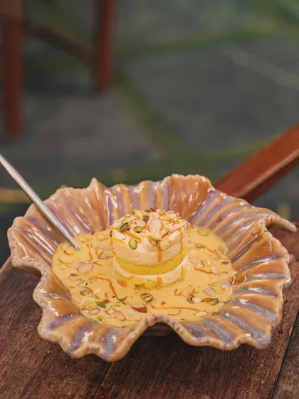 A beautiful layered Indian dessert, possibly a rasmalai tres leches, served in a unique, leaf-shaped bowl. The soft lighting highlights the creamy texture and nut garnish.