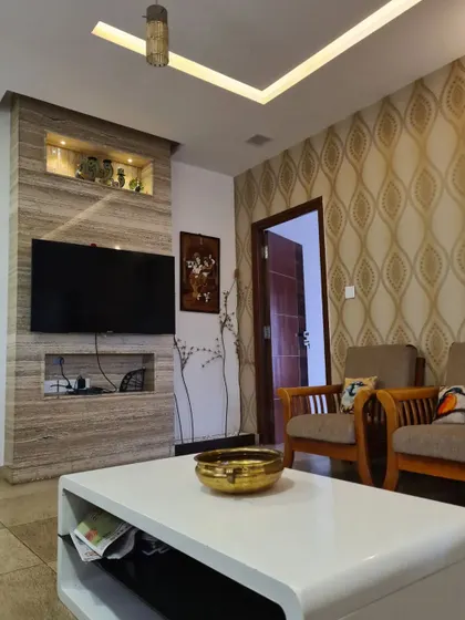 This living room showcases a custom TV unit clad in travertine stone, featuring built-in niches for electronics and decor. The design is complemented by a patterned wallpaper accent wall and a tray ceiling with recessed lighting, creating a warm and textured environment.