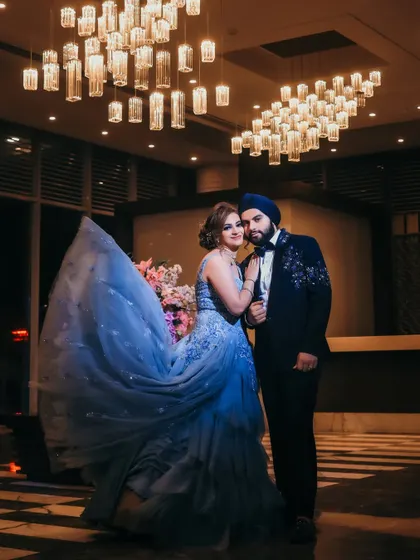 A dramatic, wide-angle shot of the couple, with the bride's blue gown flowing beautifully. The modern chandelier and moody lighting create a cinematic and high-fashion feel.