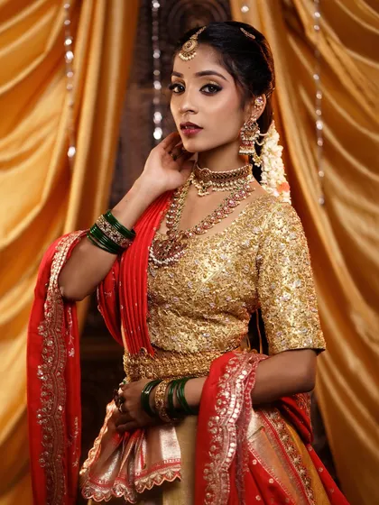 A full-view portrait of a bride in a red and gold half-saree. The makeup and styling are perfectly suited for pre-wedding ceremonies or a traditional engagement.