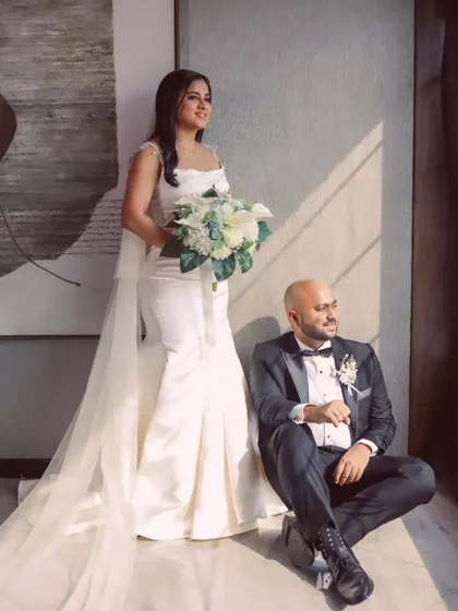 A modern, chic portrait. The bride stands tall and elegant in her fitted gown, while her groom sits casually, creating a cool, stylish composition.