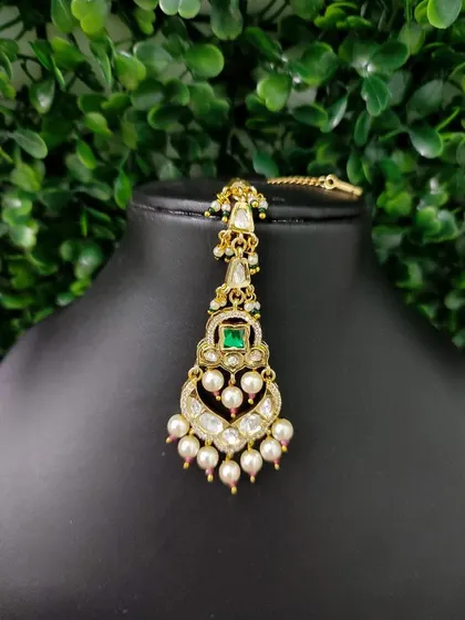 A delicate maang tikka with a heart-shaped design, featuring a central emerald stone and pearl drops. A lovely choice for pre-wedding functions.
