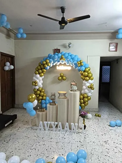 A home birthday setup with a circular balloon arch in blue, white, and gold. The arrangement includes white plinths of varying heights.